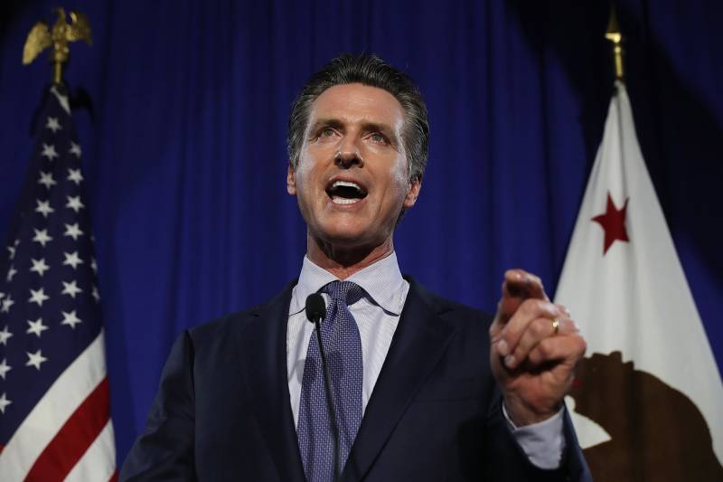 A photo of a politician in a navy blue suit with a navy and white tie stands speaking from a podium with the U.S. and California state flags behind him.