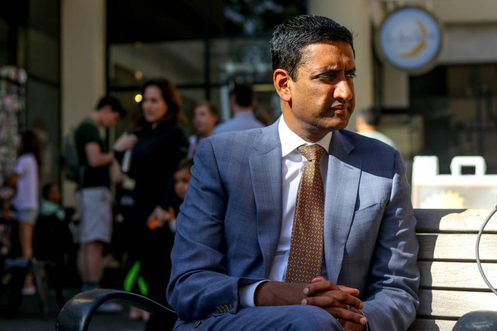 A man with dark hair and eyes wears a light blue business suit and busy orange and green tie sits on a wooden bench outside. He sits crossed-legged with his arms folded on his knee. He looks to the right of the camera. Crowds of people and children are pictured behind him.