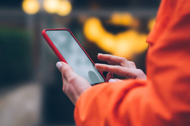 A close up of a woman's hands as she holds a smartphone and is swiping the screen. She wears an orange jacket.