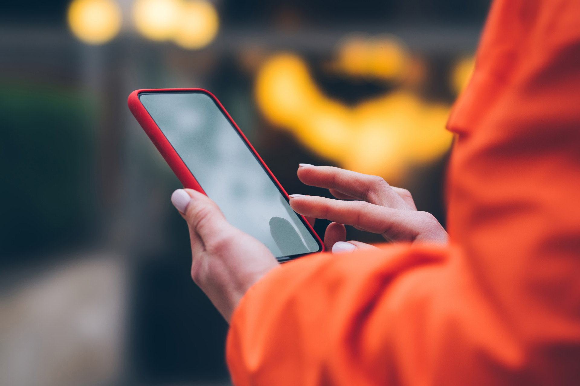 A close up of a woman's hands as she holds a smartphone and is swiping the screen. She wears an orange jacket.