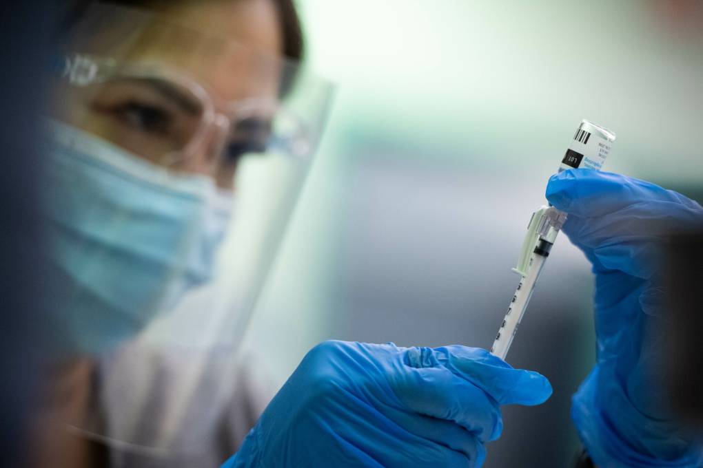 A healthcare worker wears a face shield, a blue surgical mask and goggles, and holds a needle full of vaccine up.