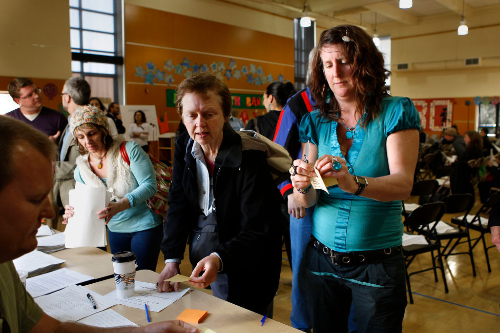 A white woman standings preparing a document, wearing a blue dress, and a man and woman beside her in a large crowded hall.