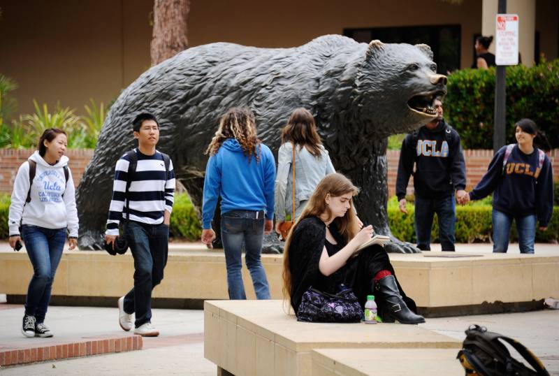 A large, bronze statue of a bear showing its teeth is displayed outdoors on a university campus. Six students in UCLA hooded sweatshirts and jeans walk around the statue on their way to class. One young woman sits on a tan, chunky bench writing on a notepad.