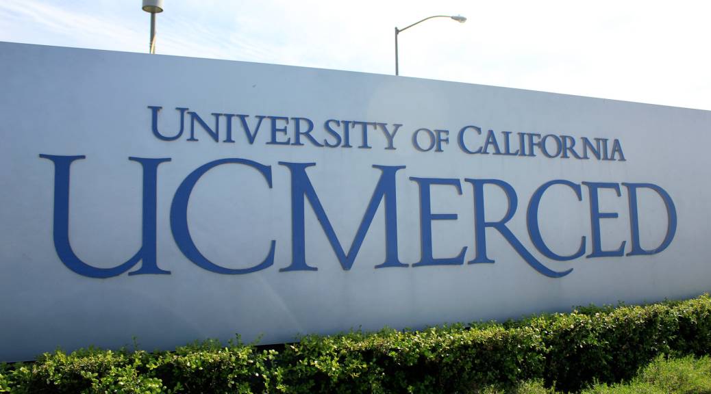 A large, concrete sign outdoors reads, "University of California UC Merced" in blue letters. Small, green bushes line the bottom of the large sign.