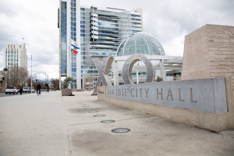 Outside of a tan and gray government building, a large metal sign reads, "San Jose City Hall." An art sculpture in the background depicts the large letters, "XO." A California State flag with a brown bear is waving from a flagpole in the background.