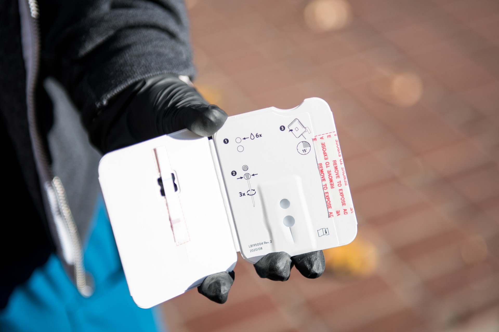 A close-up shot of a health care professional in black gloves holding a COVID-19 rapid test, which is pictured as a thick, white paper test with holes for a cotton swab to rest inside.