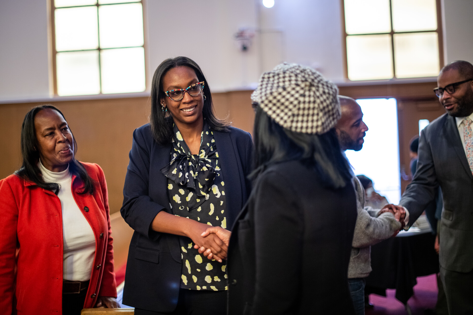A Black woman wearing glasses, a navy blue jacket and yellow and black designed shirt, shakes hands with a Black woman wearing a patterned hat and black shirt in a building with other people in the background.