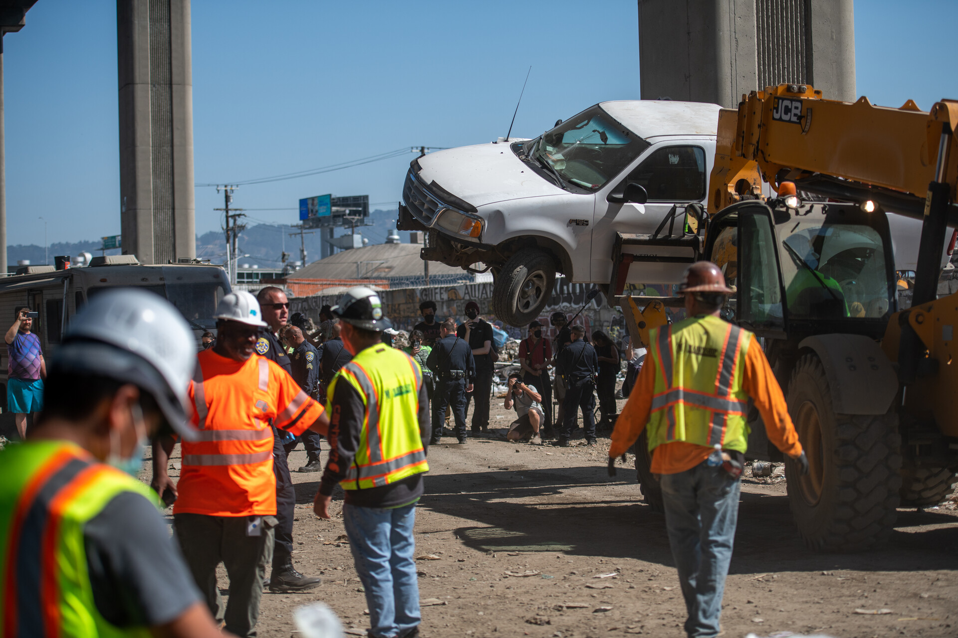Men in orange and yellow work clothes and white hardhats clear a homeless encampment using large machinery. A white pickup truck is seen being hoisted into the air and hauled off.