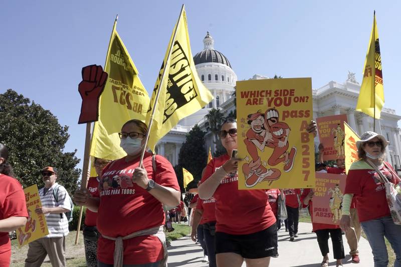 A large group of protesters wearing red T-shirts march around California's Capitol building in Sacramento. Many are carrying yellow signs with red writing. One sign reads, "Which Side Are You On? Pass AB257."