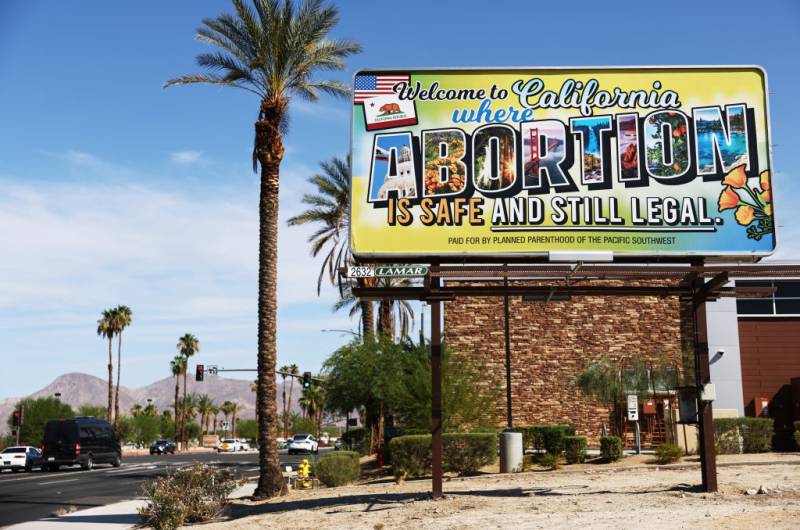 A colorful billboard in front of palm trees on the side of a road reads, "Welcome to California, where abortion is safe and still legal."