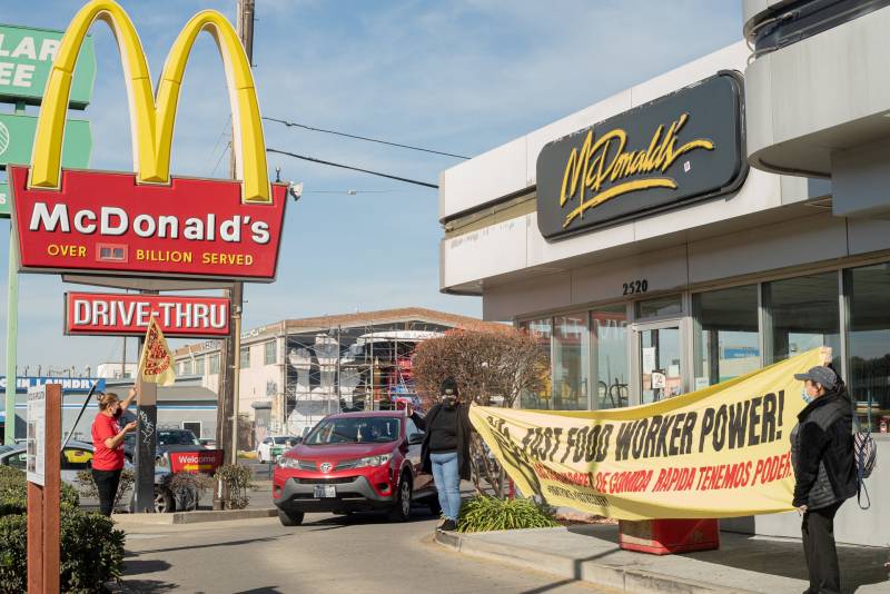 A McDonald's drive-thru is pictured, where several protesters are seen holding a large yellow banner with black and red writing that reads, "Fast Food Worker Power!" A maroon SUV is pulling out of the drive-thru.