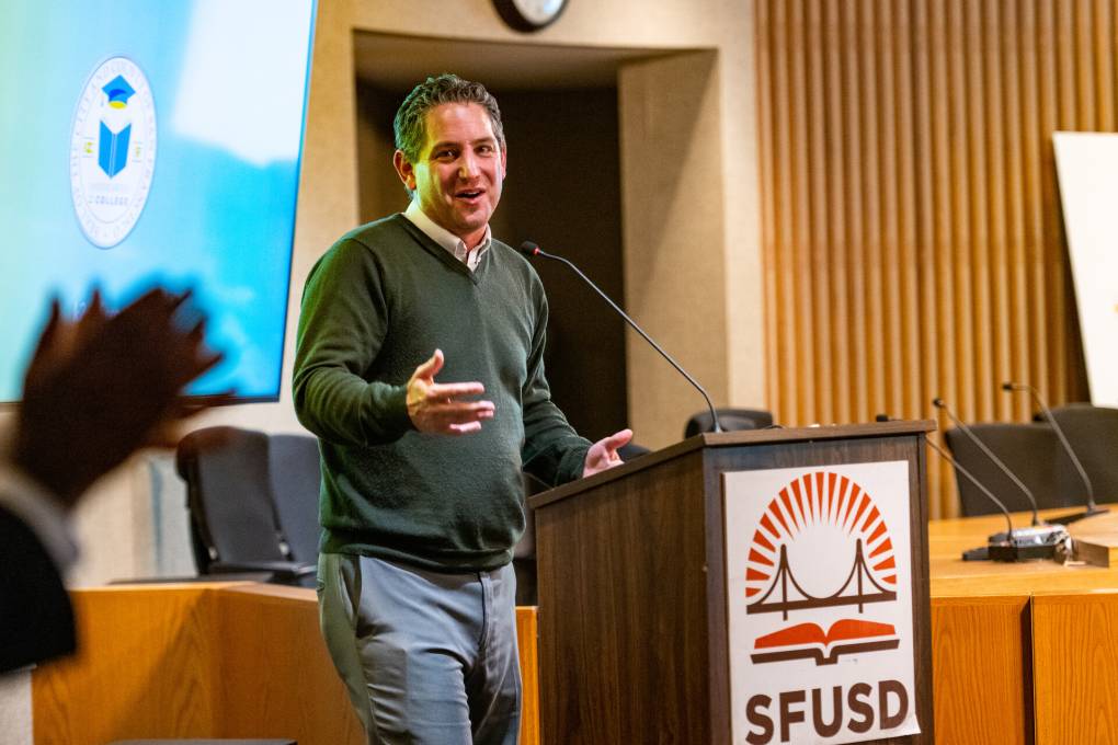 A white man speaks to the audience from behind a dais with an "SFUSD" emblem on it.