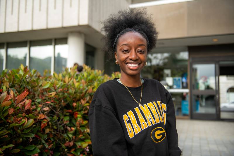 An African American girl smiles at the camera with a school behind her.