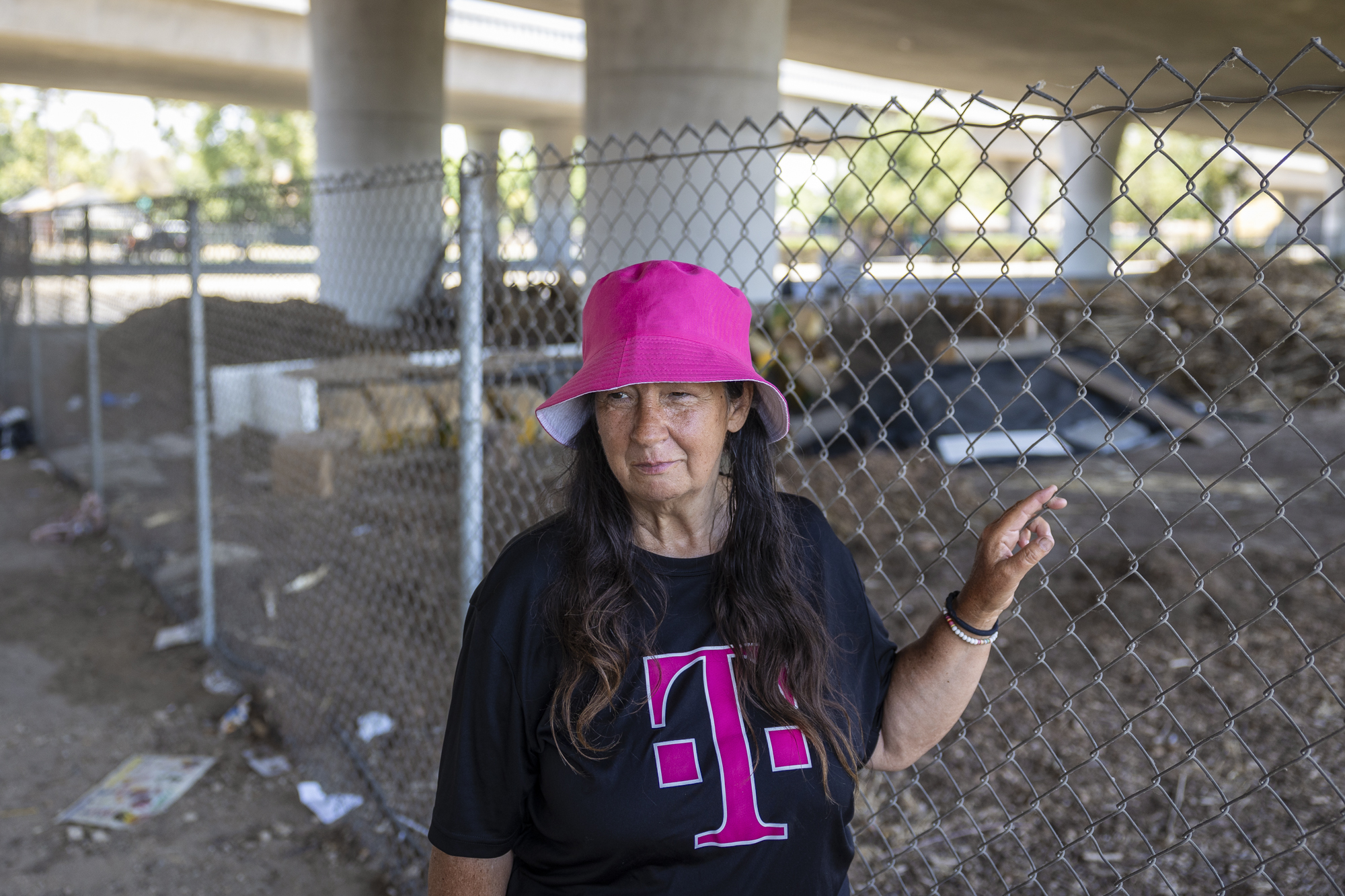 A woman in a pink hat leans on a chainlink fence under a freeway overpass.