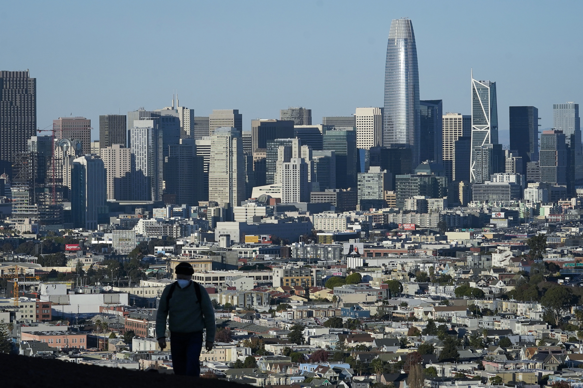 A person walks up a hill with the San Francisco skyline in the background.