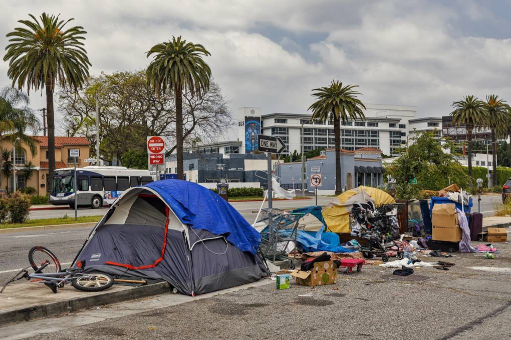 Tents line a paved road near a sidewalk. A bicycle is on its side in front of a gray and navy tent with a blue tarp covering its roof. Scattered belongings and cardboard boxes cover the spaces between each tent. A city bus is seen driving in the background.