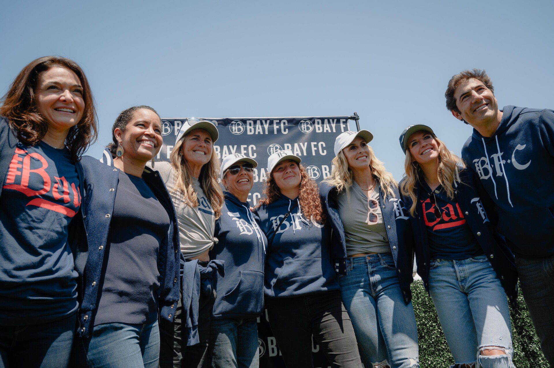 A row of people smile at the camera, they are all wearing items of clothing with the logo for a women's soccer team called "Bay FC."