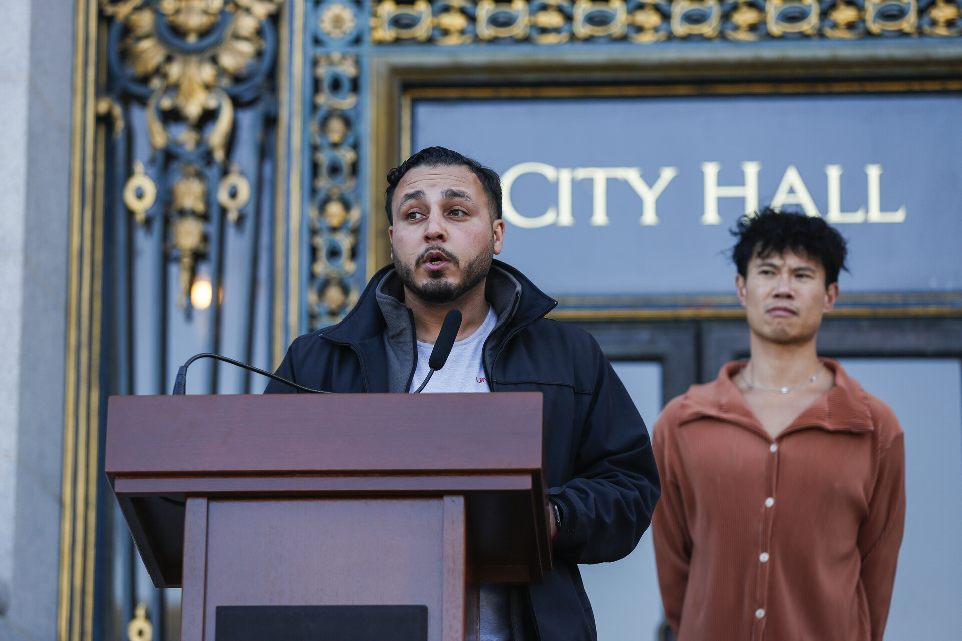 A man wearing a dark hooded sweatshirt stands at a podium with another man standing to his right wearing a reddish shirt in front of a building that has a sign reading "City Hall."