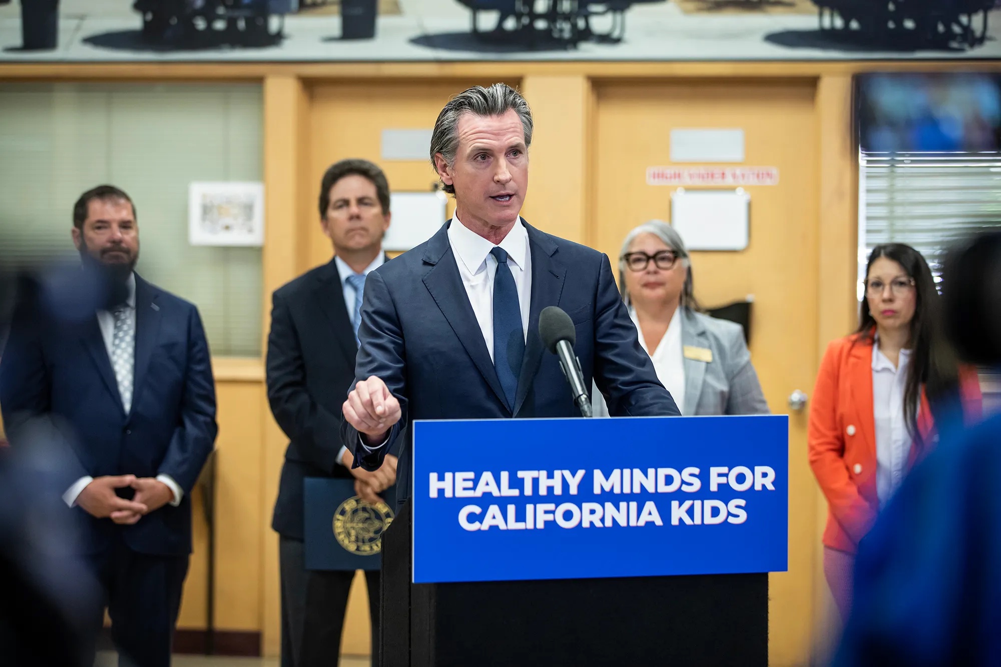 A white middle-aged man in a blue suit and blue tie speaks behind a dais that says "Healthy Minds For California Kids" surrounded by people.