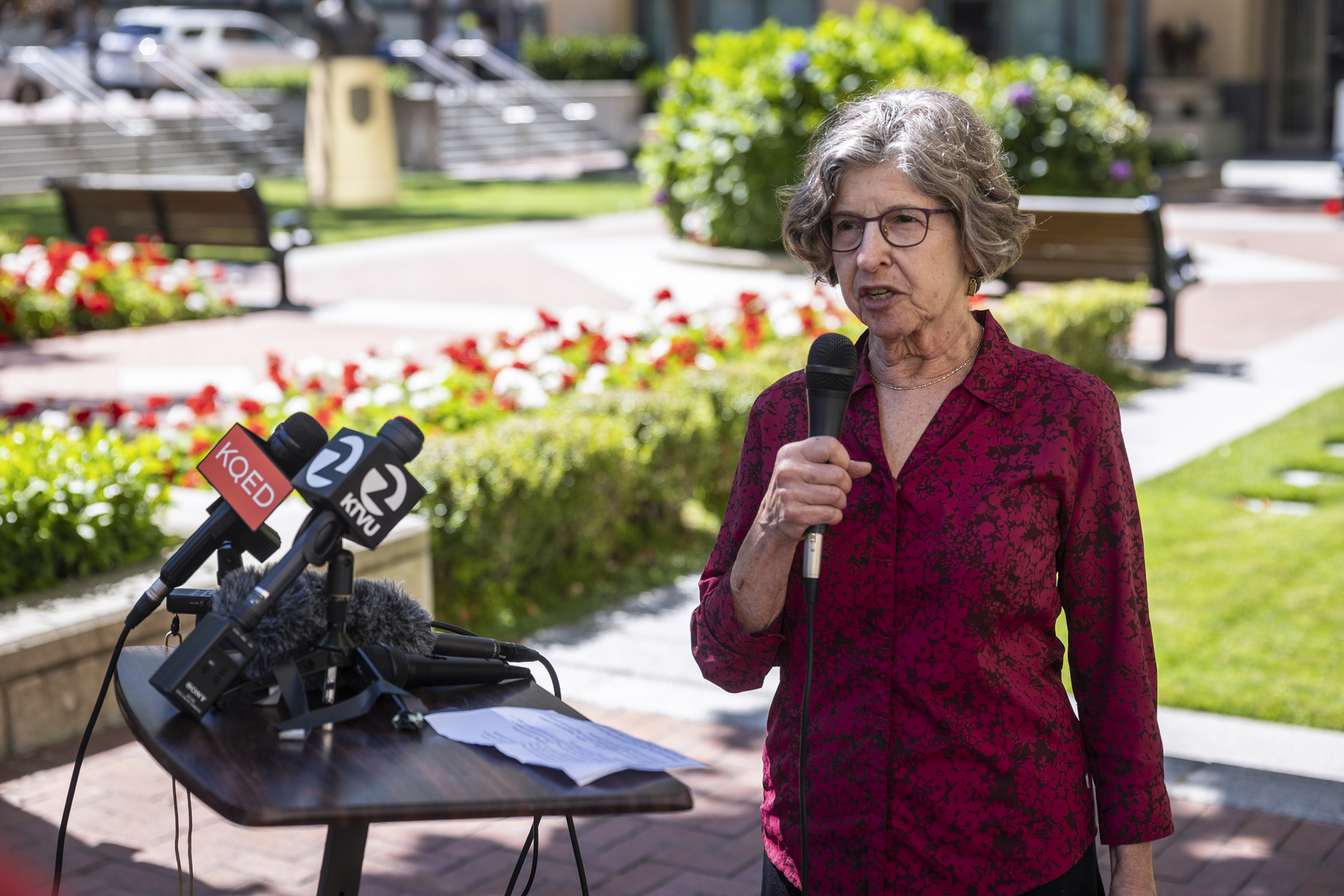 A person with glasses speaks into a microphone in an outdoor setting.