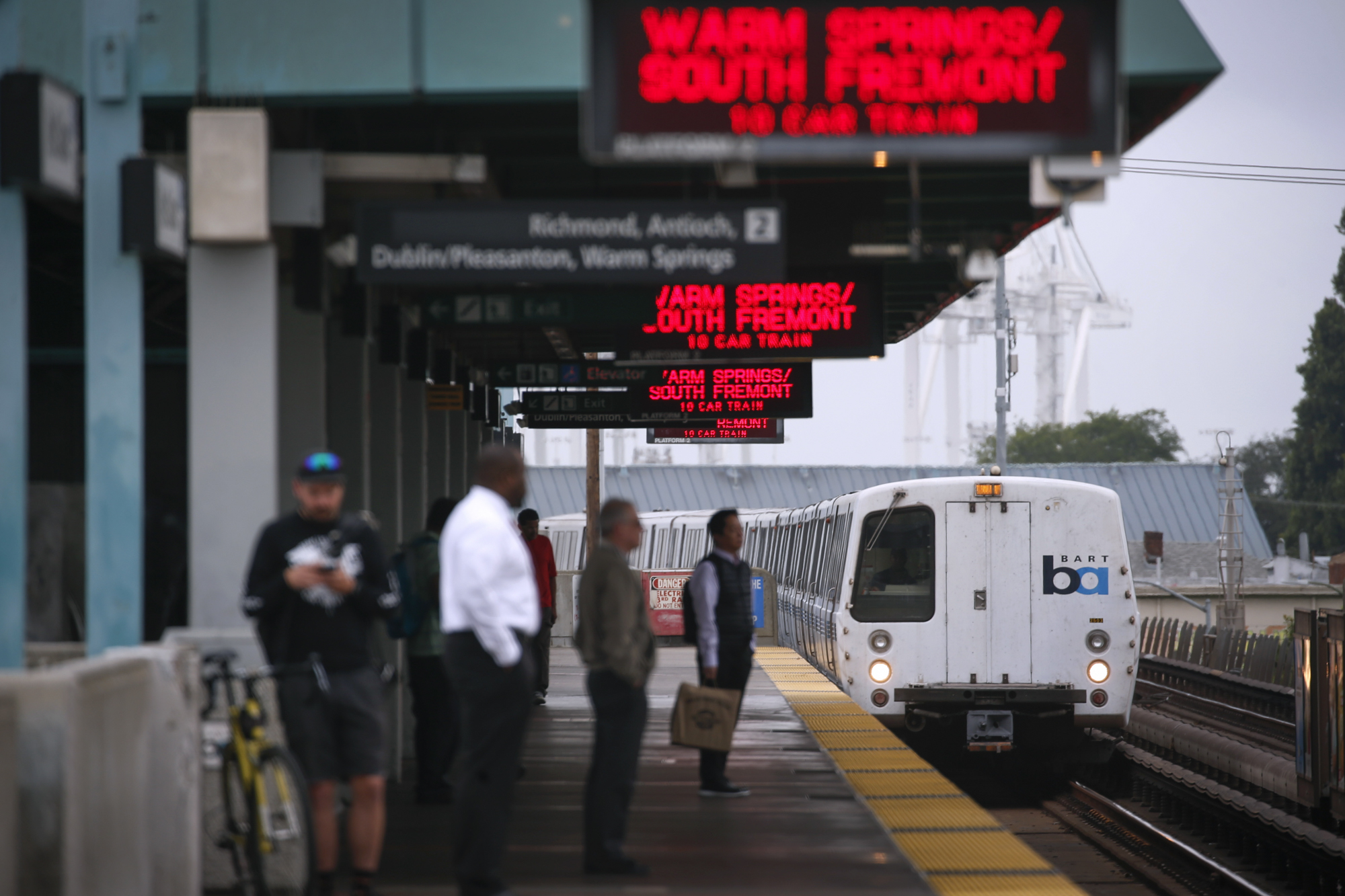 A train car pulls into an outdoor station with several passengers waiting on the platform.