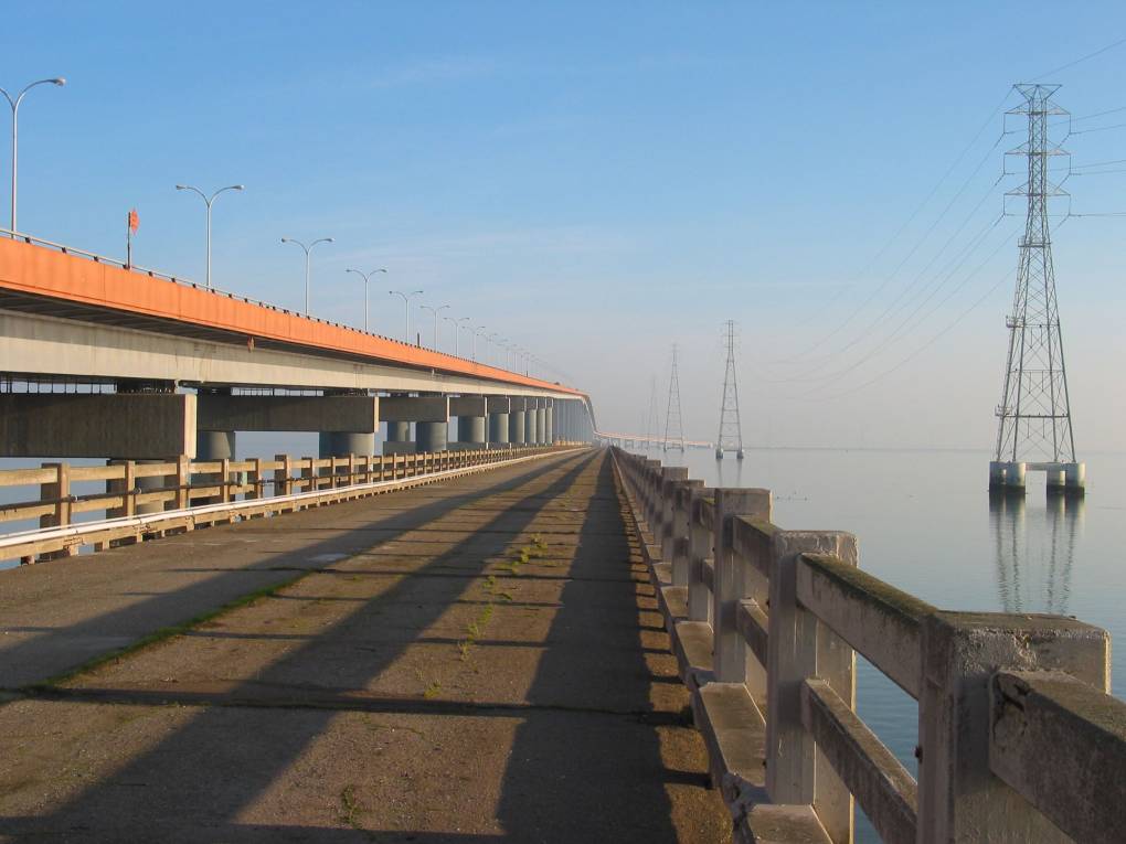 Looking down an old section of a now unused bridge that runs off into the distance and terminates just before the horizon line. To the left of it is the new bridge that replaced it. Both bridges run over calm, reflective blue water.