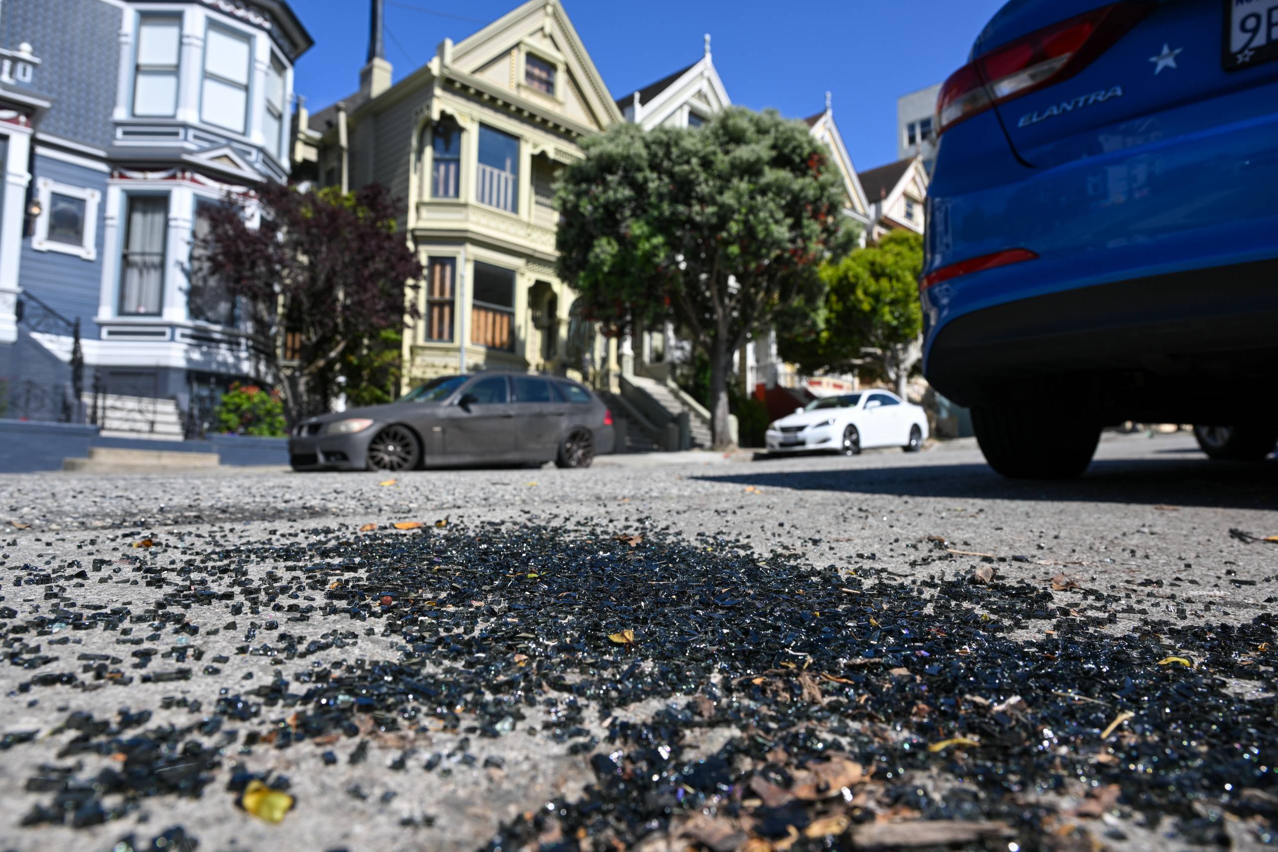 A street in San Francisco lined with Victorian homes and cars. The camera is angled from a lower angle to focus on shards of shattered glass on the floor.