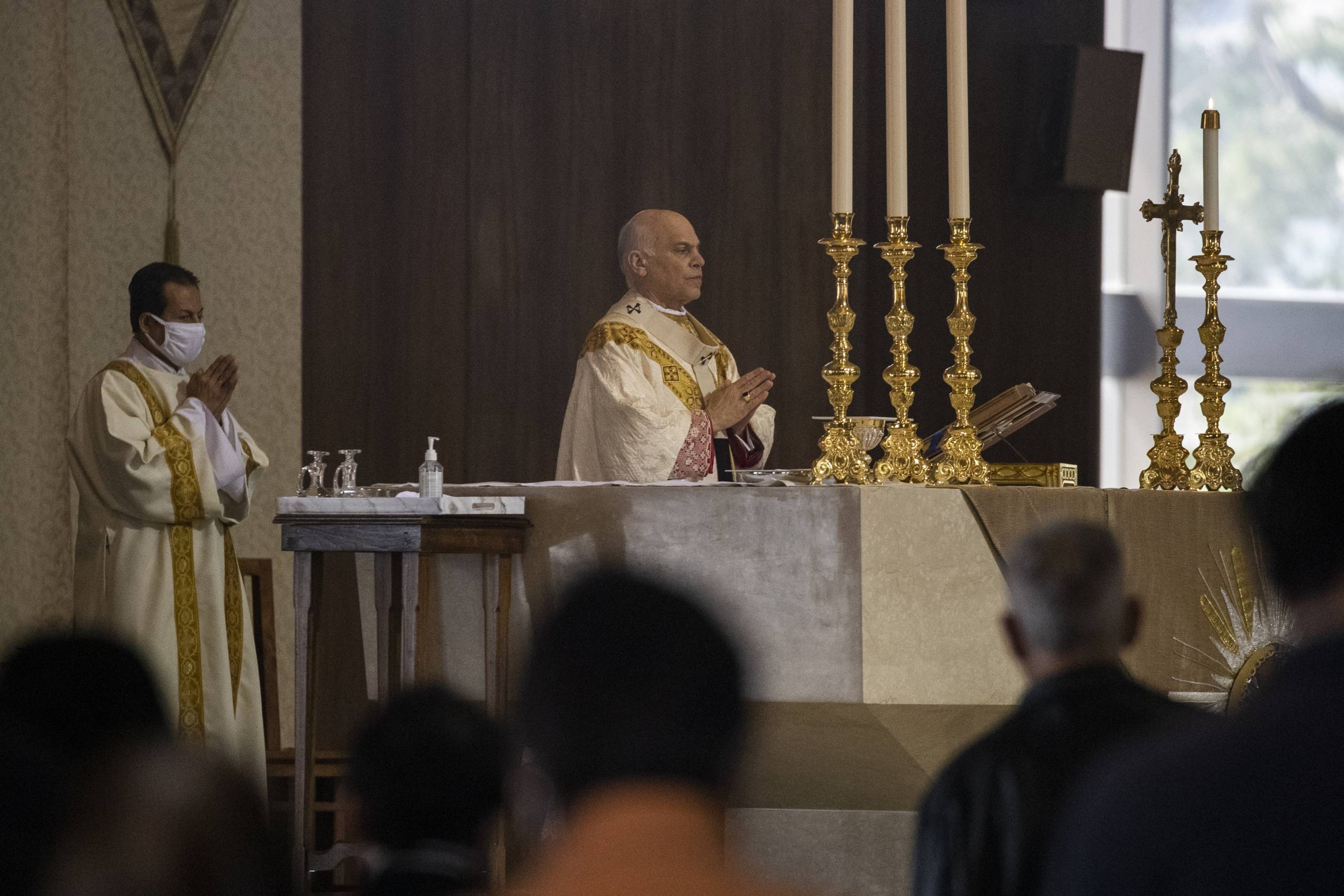 Archbishop Salvatore Cordileone wears his robes in front of the altar, facing the faithful. At the altar there are many candles. Deacons stand behind Cordileone in prayer.