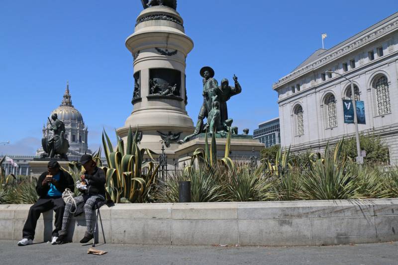 Two people sit in front of a statue and monument while they hold food and plastic bags in their laps.