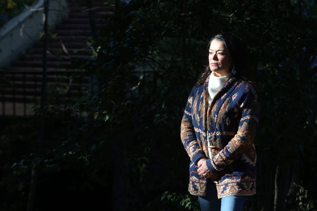 A woman stand in the shadows. Her face and body are illuminated by a spotlight. She has long, brown hair and wears a blue and tan-print jacket and jeans. She looks somber.