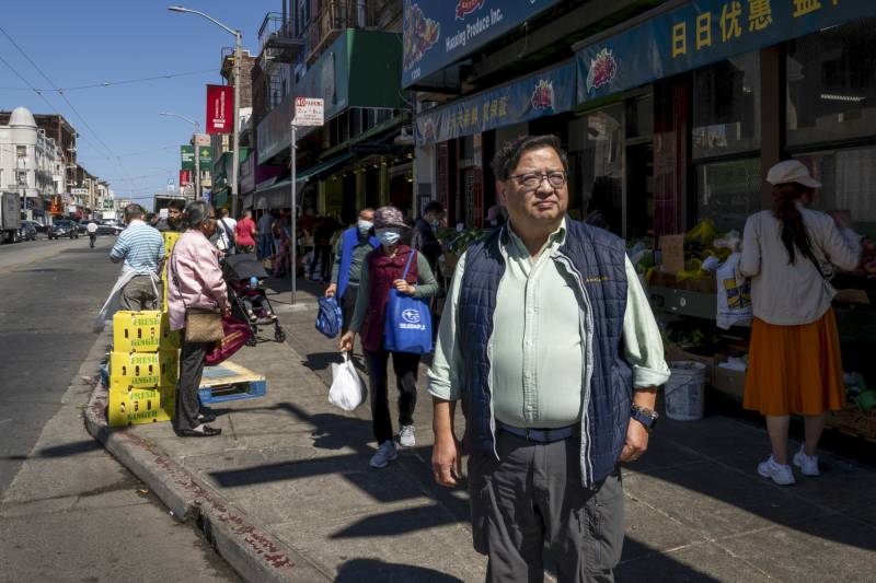 A person with glasses is photographed as other people pass on a busy city sidewalk.