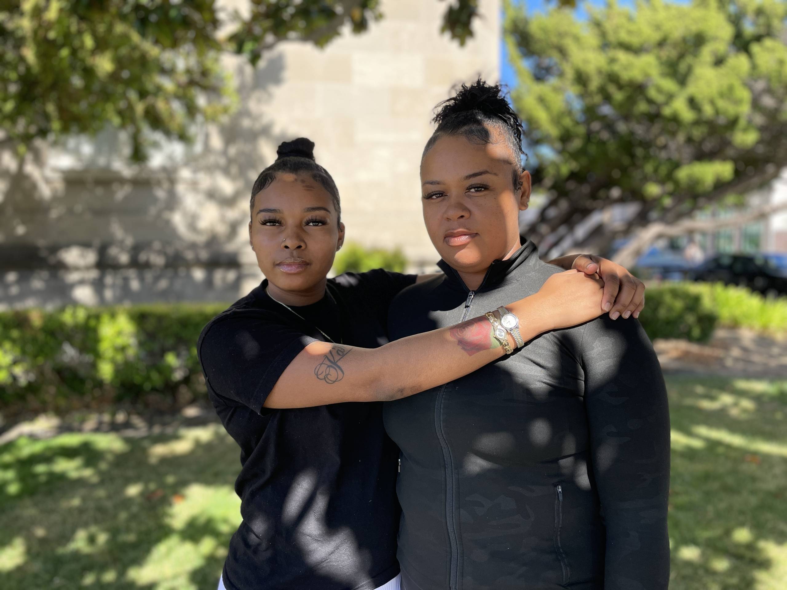Two people with their hair in buns stand together looking at the camera in an outdoor setting.