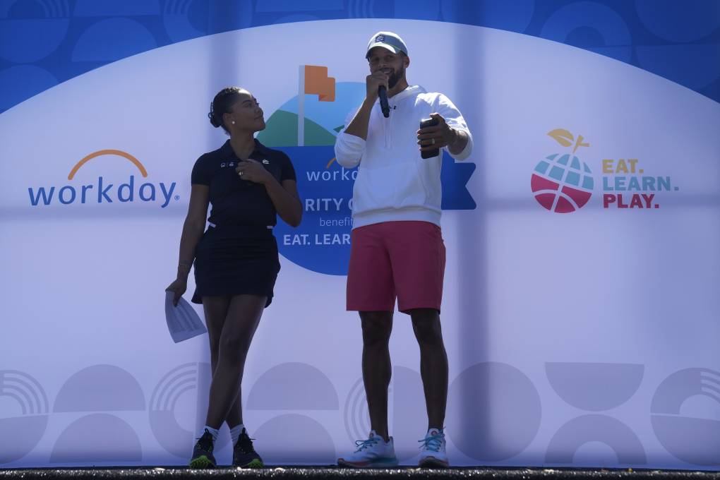 A man and a woman stand on a stage speaking with a huge banner behind the that says, "Eat. Learn. Play."