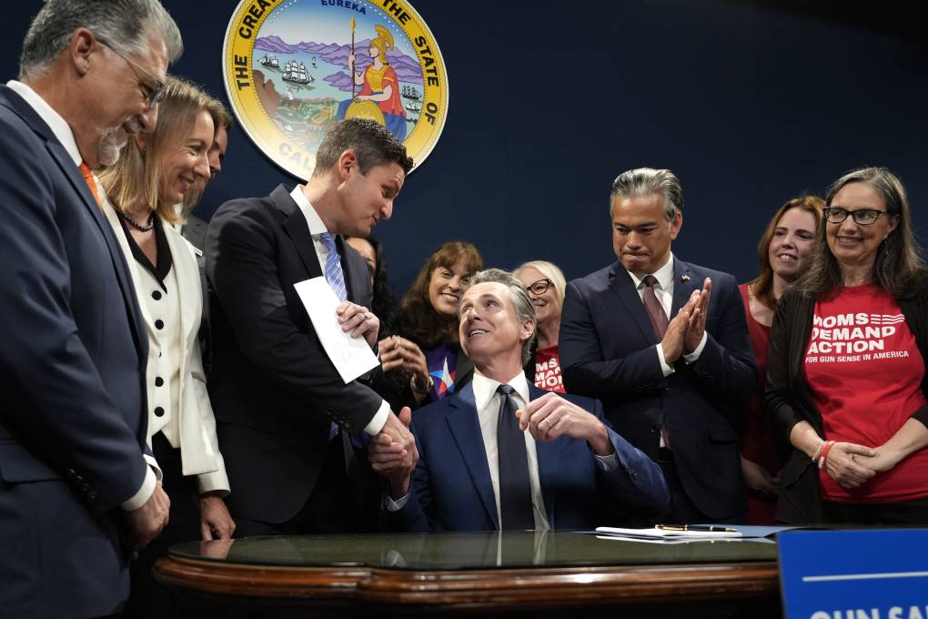 People dressed in business suits and dresses stand around a man in a business suit who looks up at a man to shake his hand.