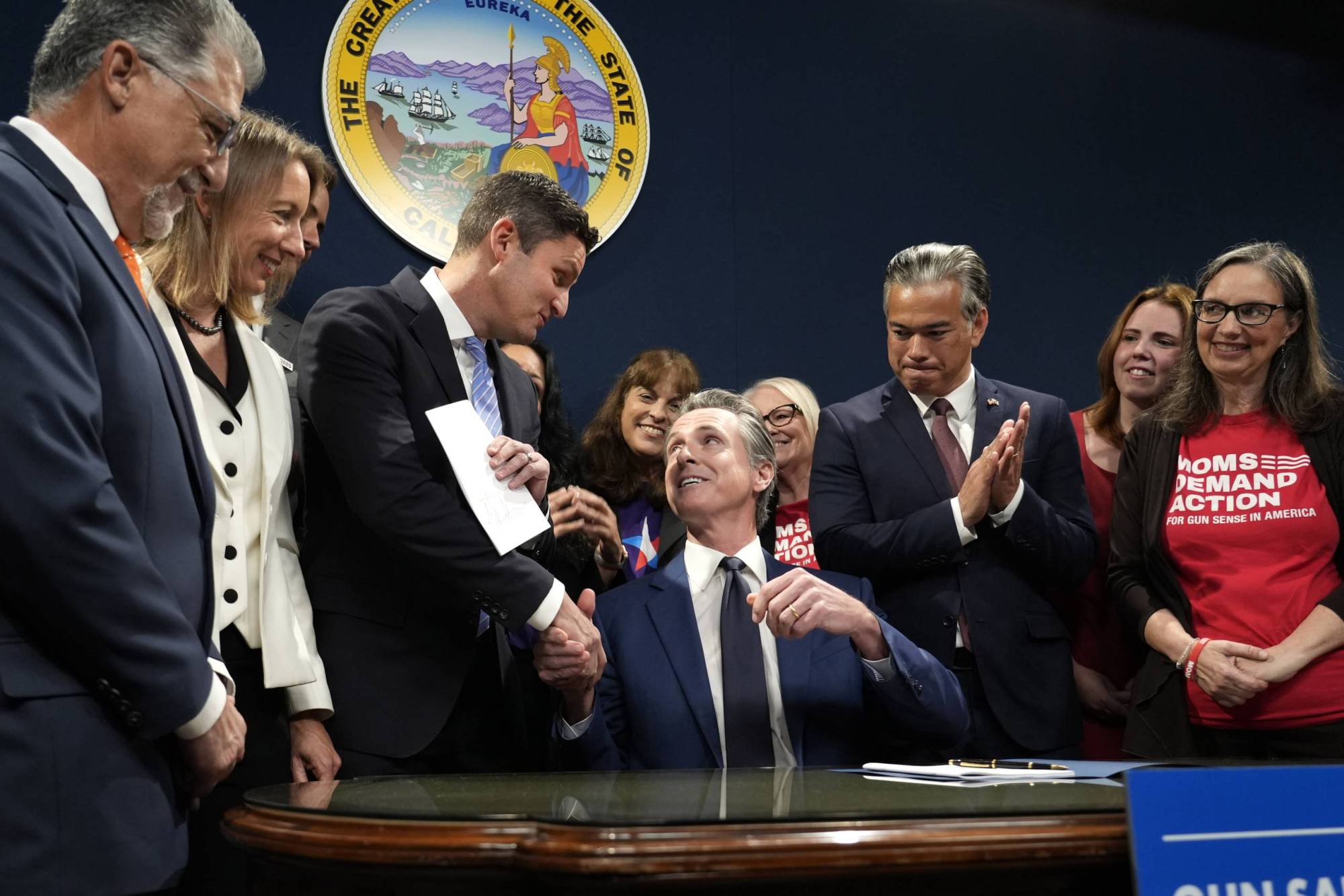 People dressed in business suits and dresses stand around a man in a business suit who looks up at a man to shake his hand.