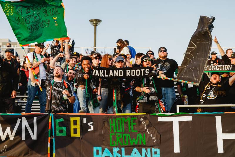 People cheer in the stands of a soccer game, waving signs that say 'Oakland Roots.'