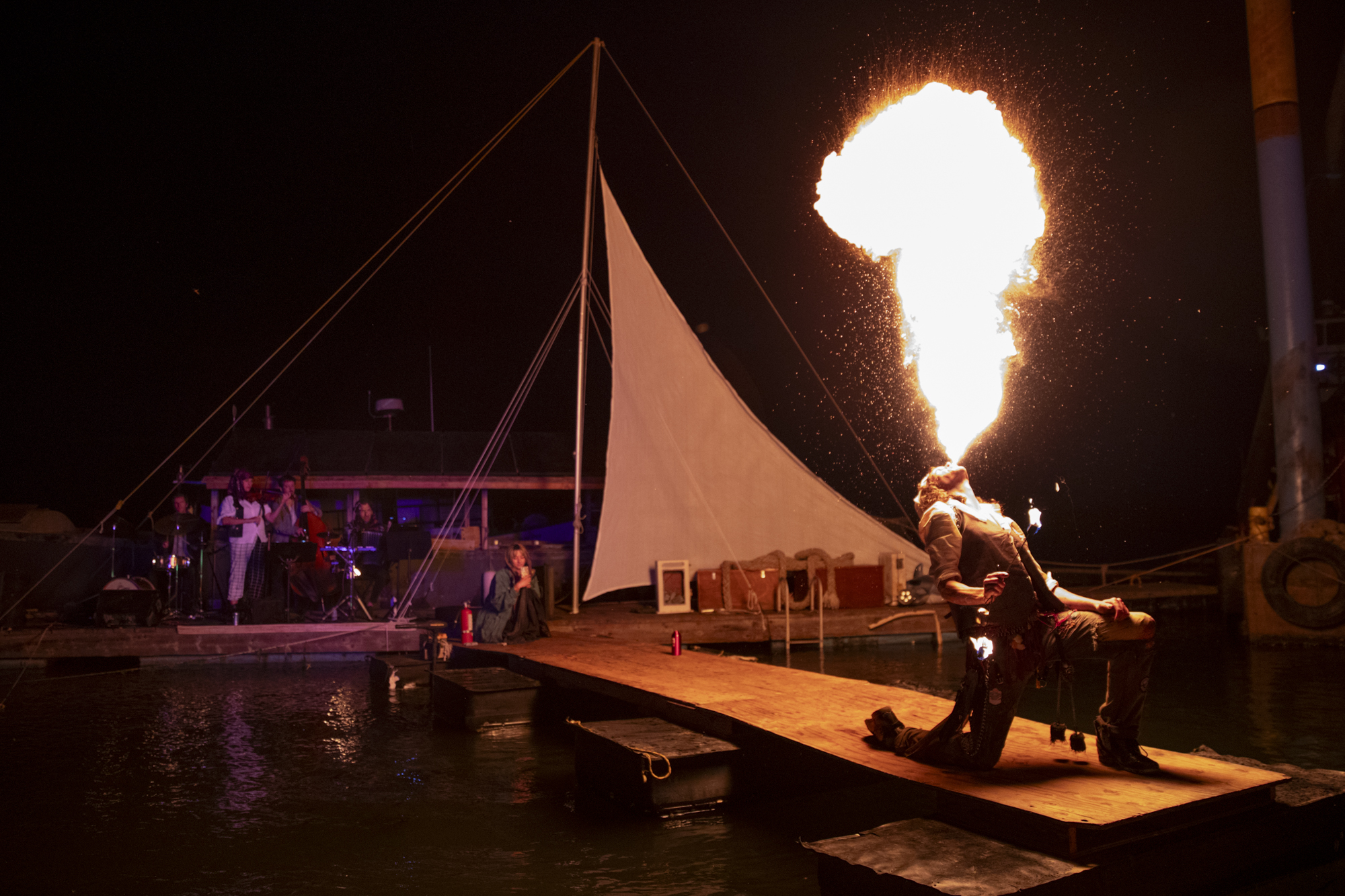 A person kneels and breathes fire at the end of a short jetty.