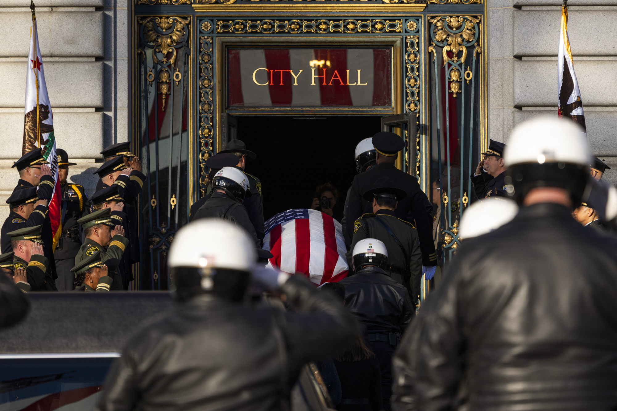 A large group of people salute a casket draped in an American flag as it enters a building with the words "City Hall" over the door.
