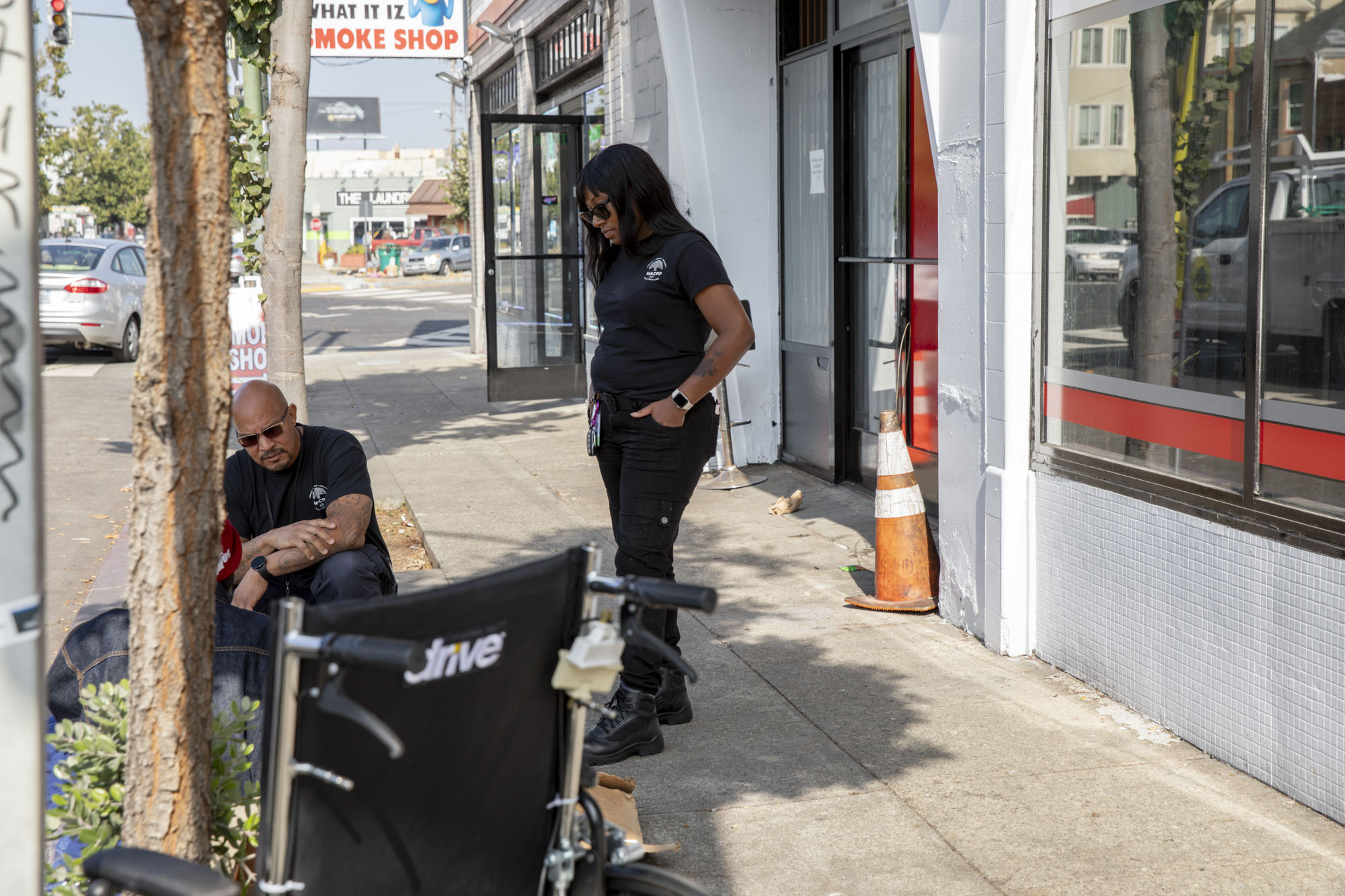 Two people en black shirts engage with a third unseen person sitting on a city sidewalk.