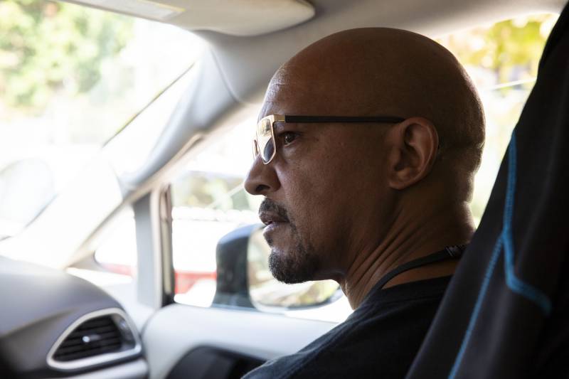 A person with a bald head looks out the driver's side window of a car from the passenger seat.