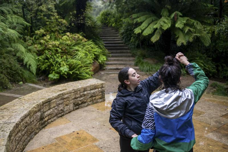 Two people hold each other, appearing to be dancing, in a wooded area.