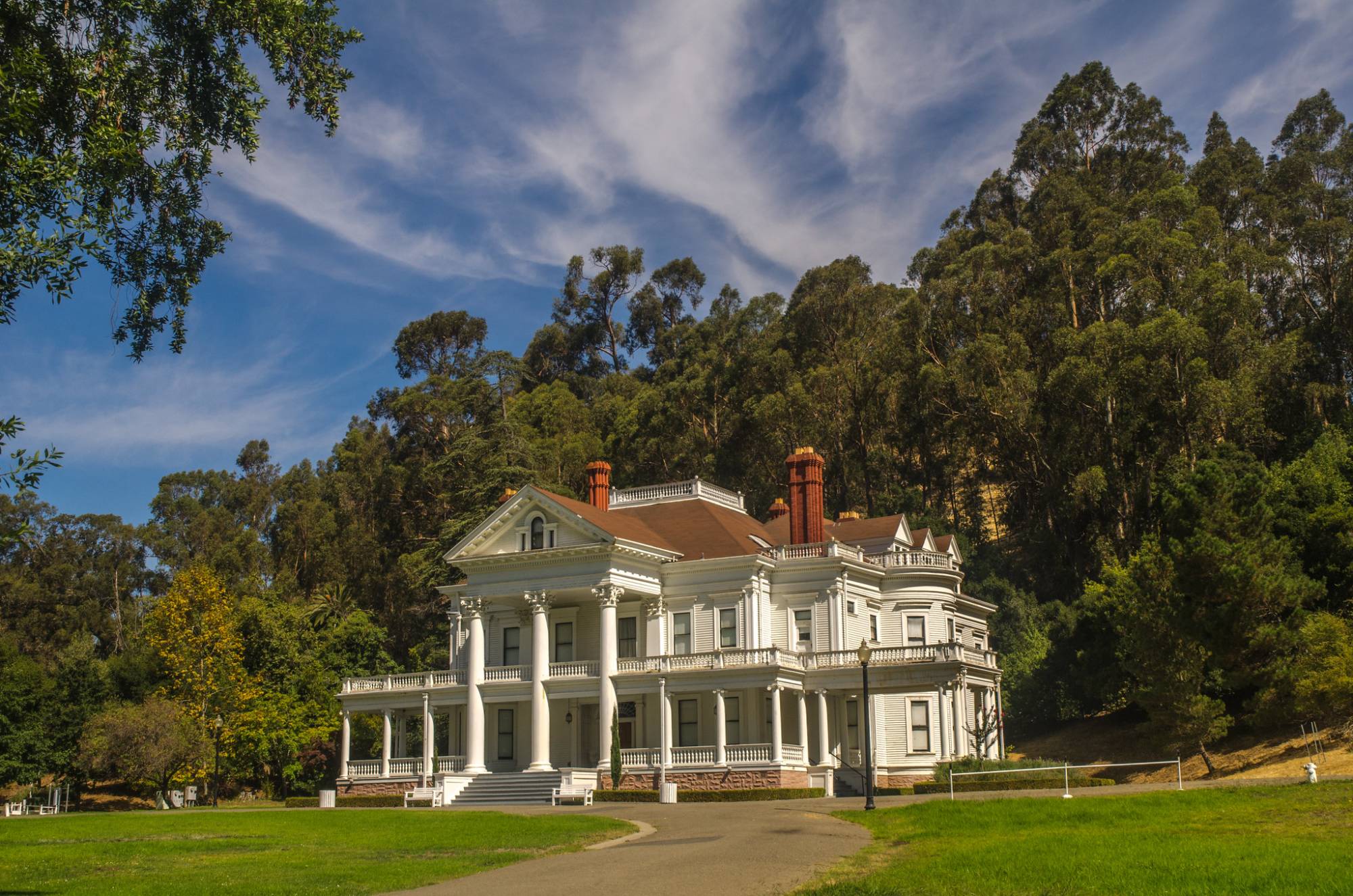 A large neoclassical white house sits behind a large lawn and in front of a stand of trees.