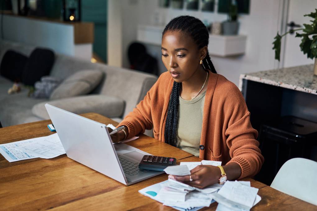 A Black woman wearing a tan sweater sits at a desk holding a piece of paper in one hand and staring at a laptop.