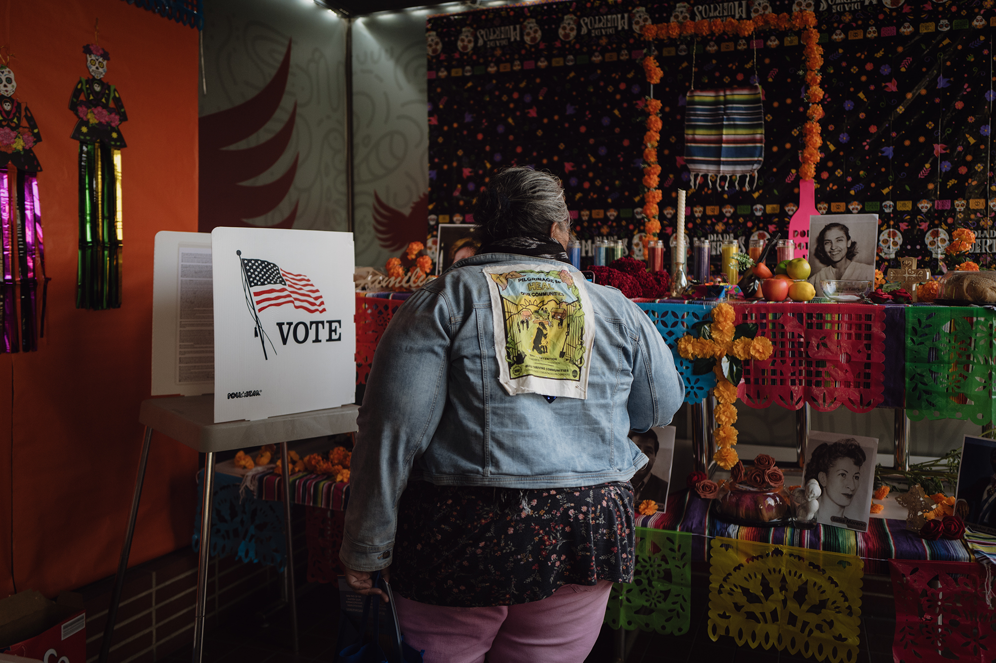 A woman has her back turned showing a jean jacket with an image in the middle, to the left of a sign with an American flag that reads "Vote."