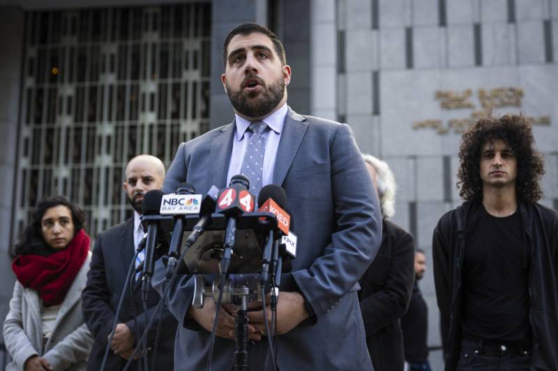 A person with a beard and wearing a suit speaks at a bank of microphones in front of a large building.