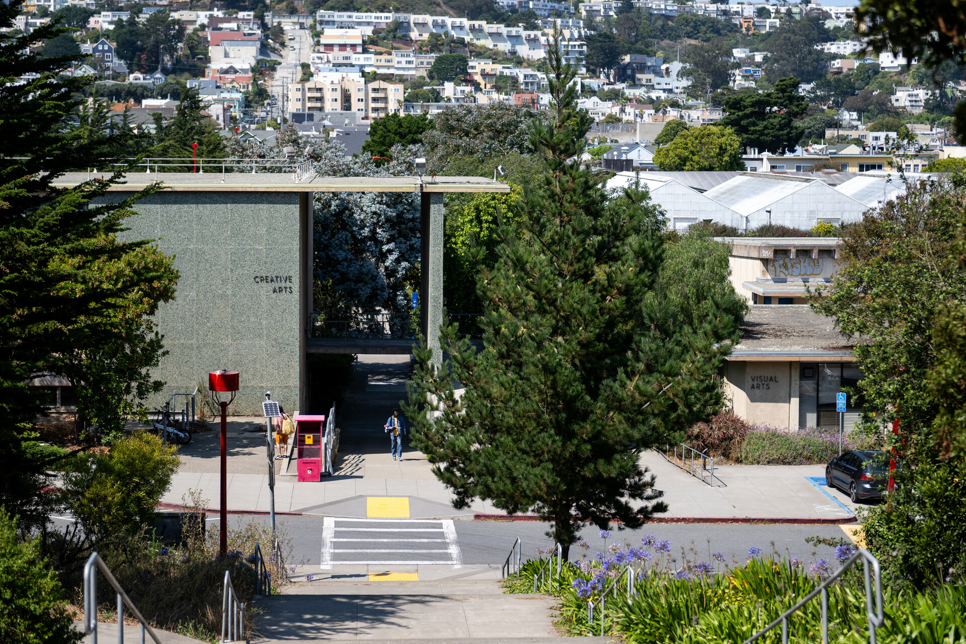 A person walks toward a building labeled "Creative Arts" on a campus with trees, sidewalks and the street in view.