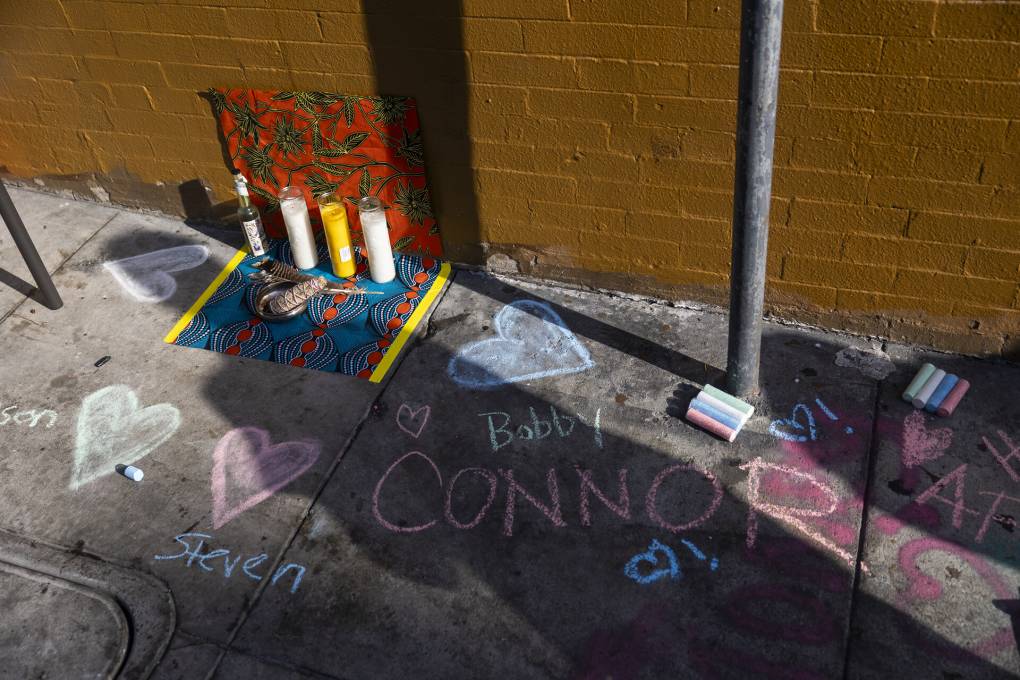 Names written in chalk appear on the sidewalk in memorial. Candles are also placed on a mat. Packs of chalk are scattered on the ground. The names "Bobby," Connor," and "Steven" can be seen.