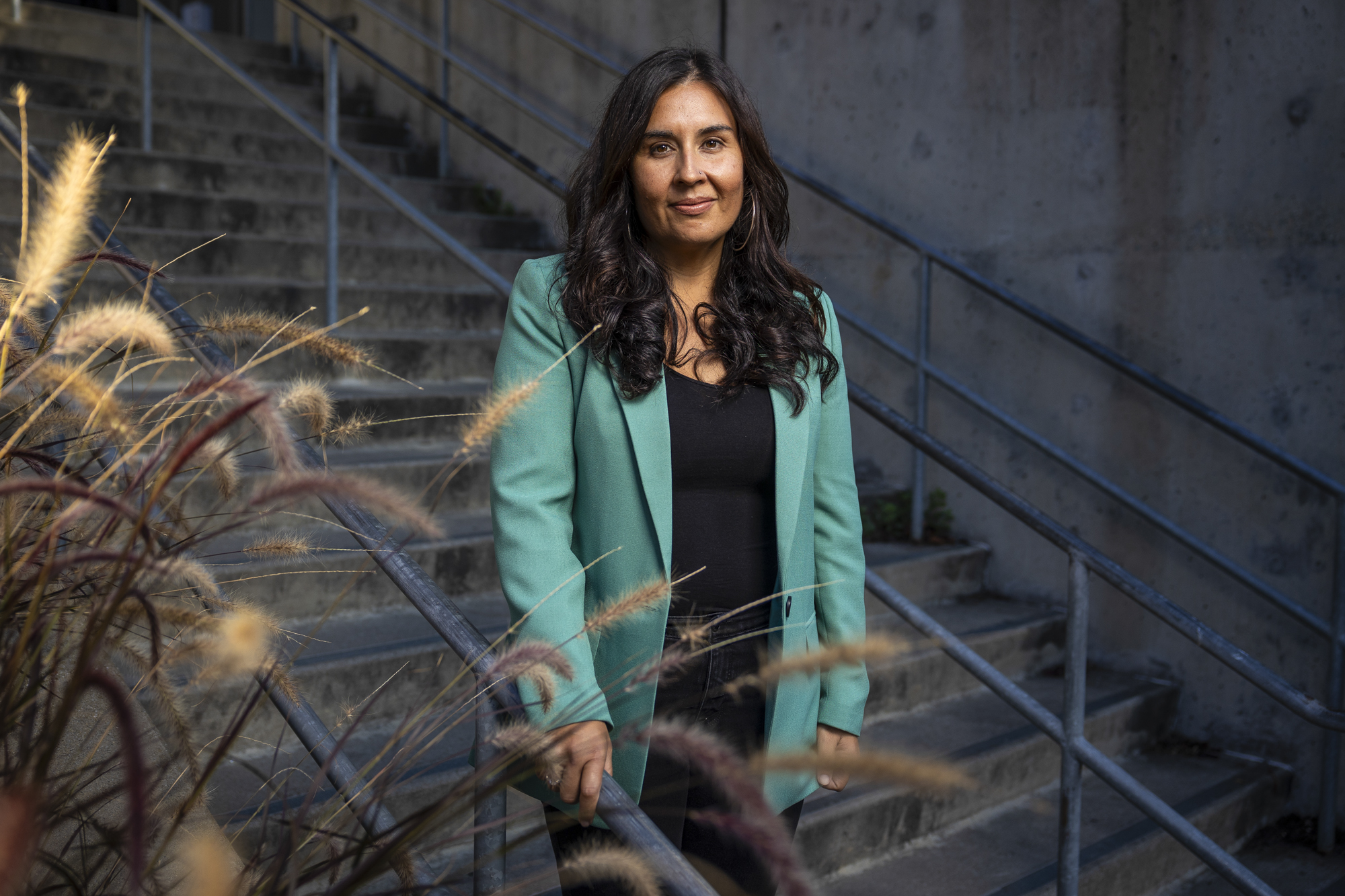 A person with long hair in a teal sport coat stands on a cement outdoor staircase and looks at the camera.