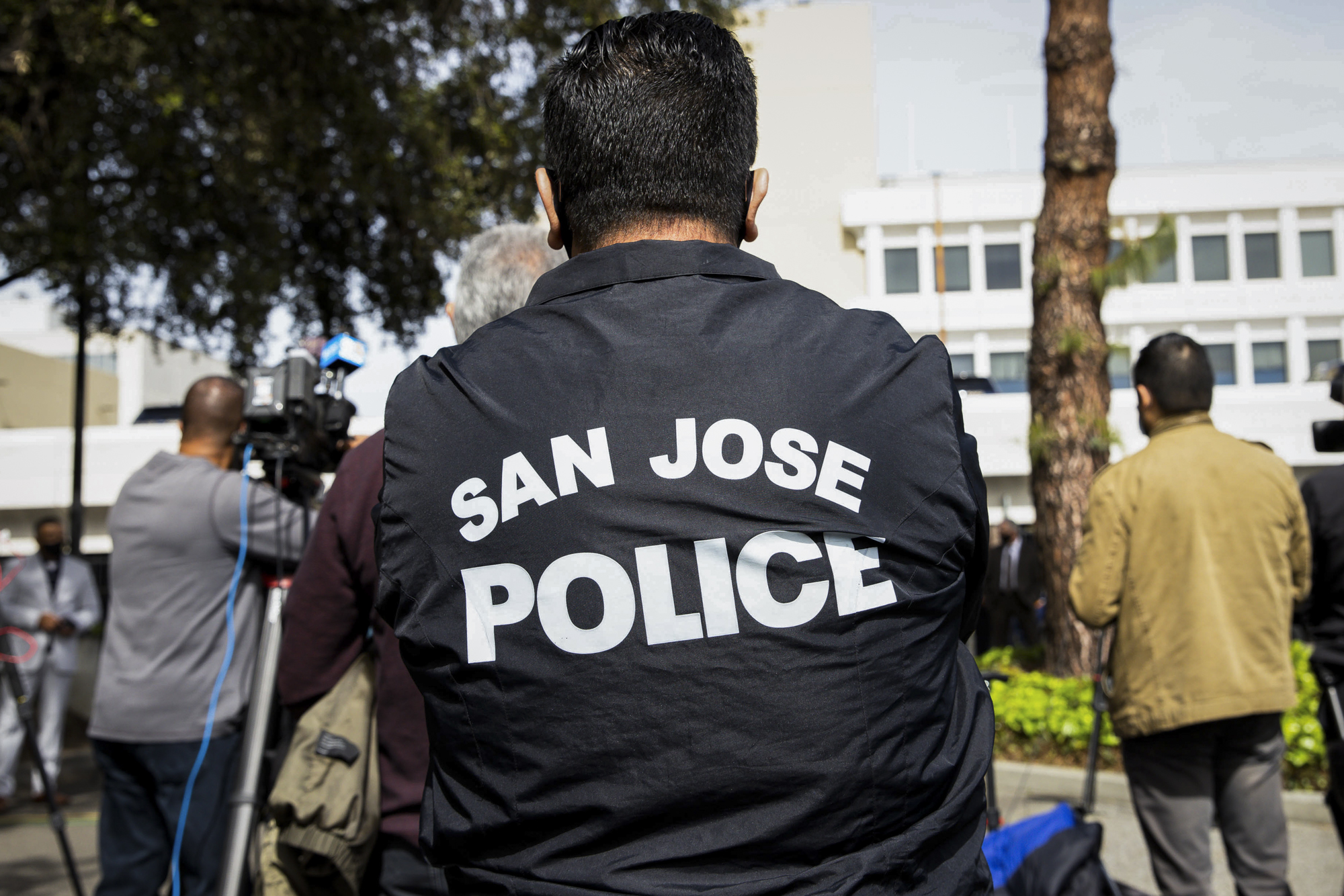 A person wears a black San Jose Police jacket in an outdoor setting.