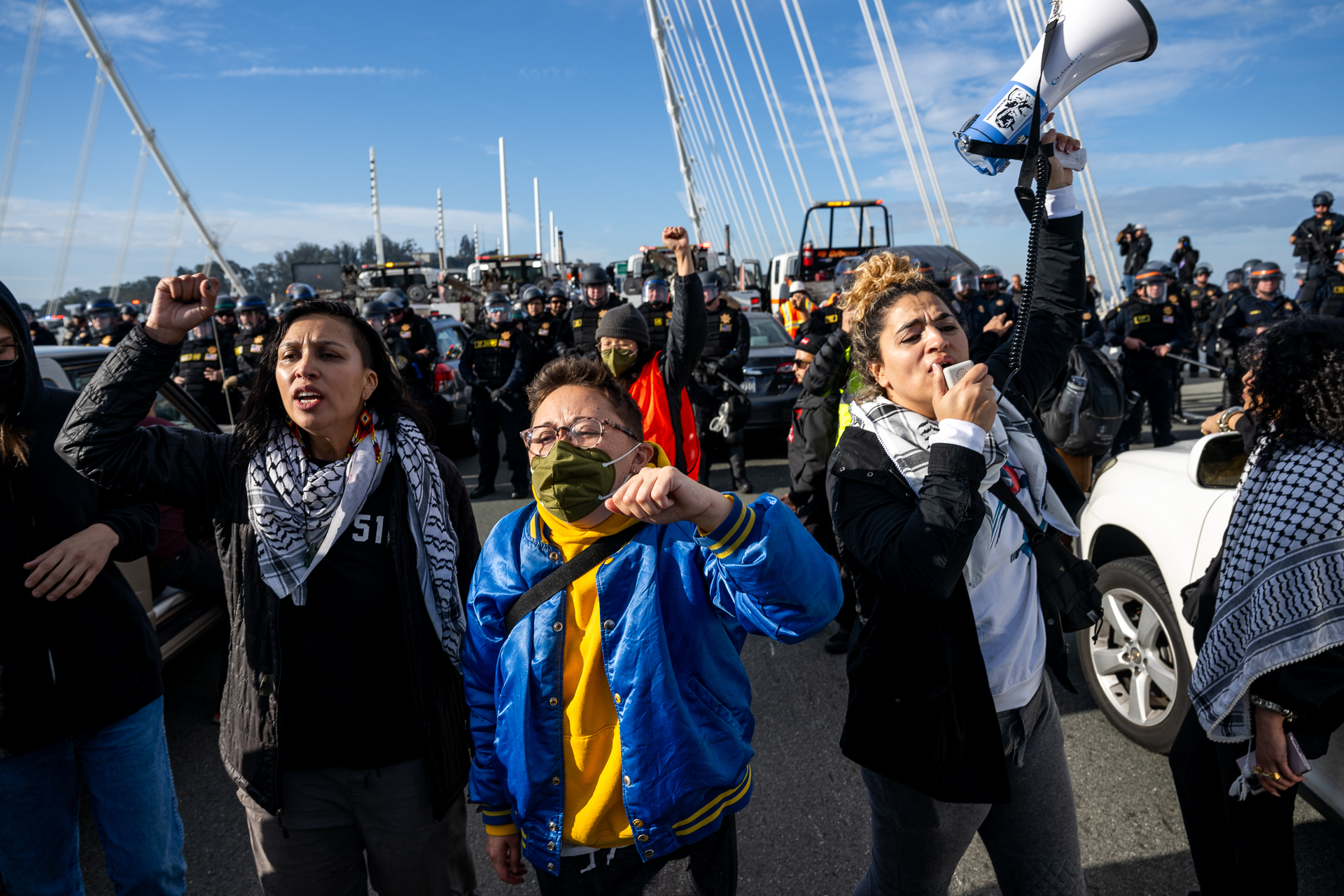 Three people chant on a bridge with law enforcement officers behind them.